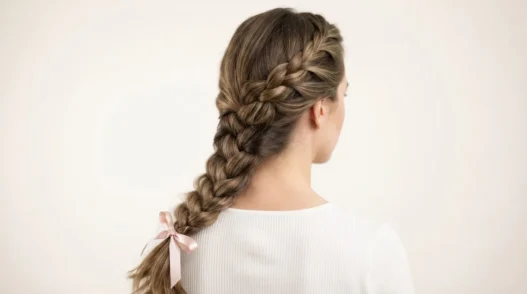 Back view of a woman with a loose three-strand side braid draped over shoulder against white backdrop in soft studio light