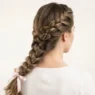 Back view of a woman with a loose three-strand side braid draped over shoulder against white backdrop in soft studio light