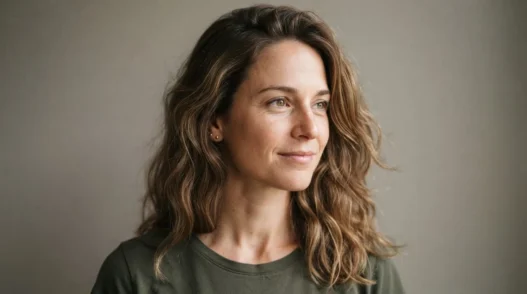 Woman with loose natural shoulder-length waves against a warm grey studio wall in soft frontal light