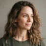 Woman with loose natural shoulder-length waves against a warm grey studio wall in soft frontal light