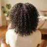 Back view of woman with damp 4B natural coils sitting on a sunlit bedroom floor with warm window light
