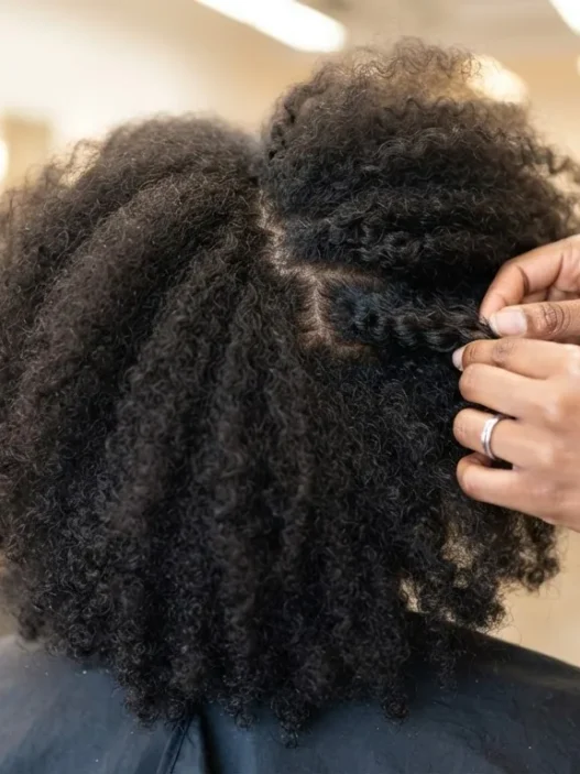 Woman in salon chair with dry natural 4C hair, hands pressing temples to demonstrate reading growth direction at root