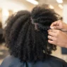 Woman in salon chair with dry natural 4C hair, hands pressing temples to demonstrate reading growth direction at root